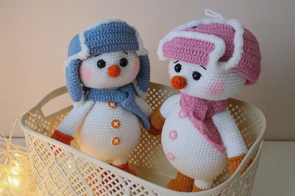 Two crocheted snowmen in ushanka hats and scarves, handmade amigurumi toys in pink and blue, standing in a basket with warm lights