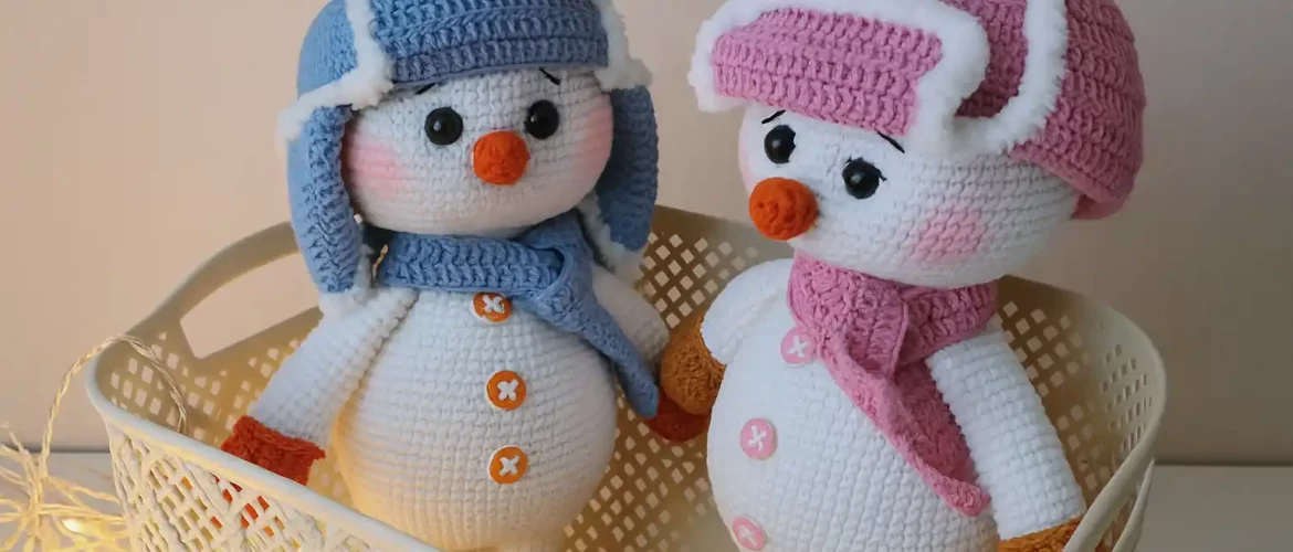 Two crocheted snowmen in ushanka hats and scarves, handmade amigurumi toys in pink and blue, standing in a basket with warm lights